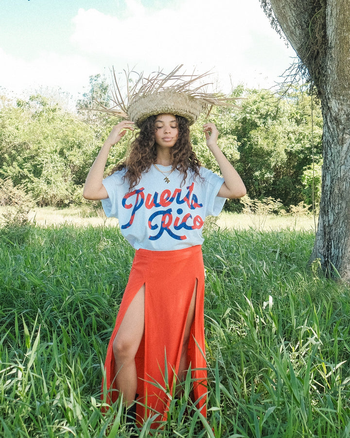 Model wearing a Puerto Rico graphic t-shirt with a straw hat and orange skirt in a grassy field