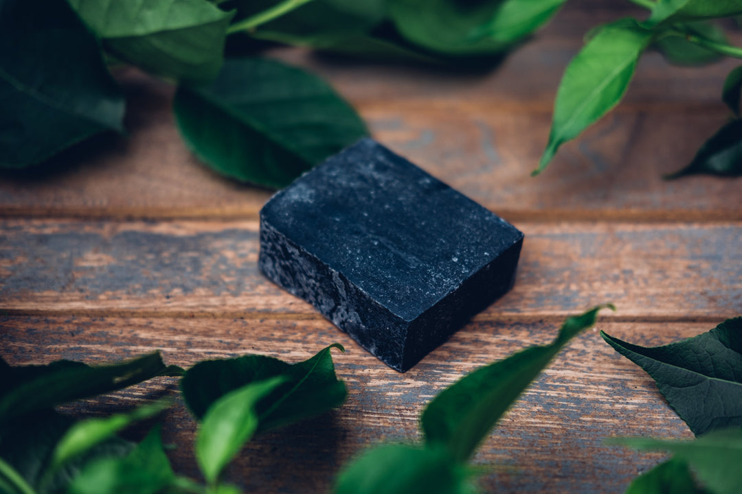 Vegan charcoal soap bar surrounded by green leaves on a wooden surface